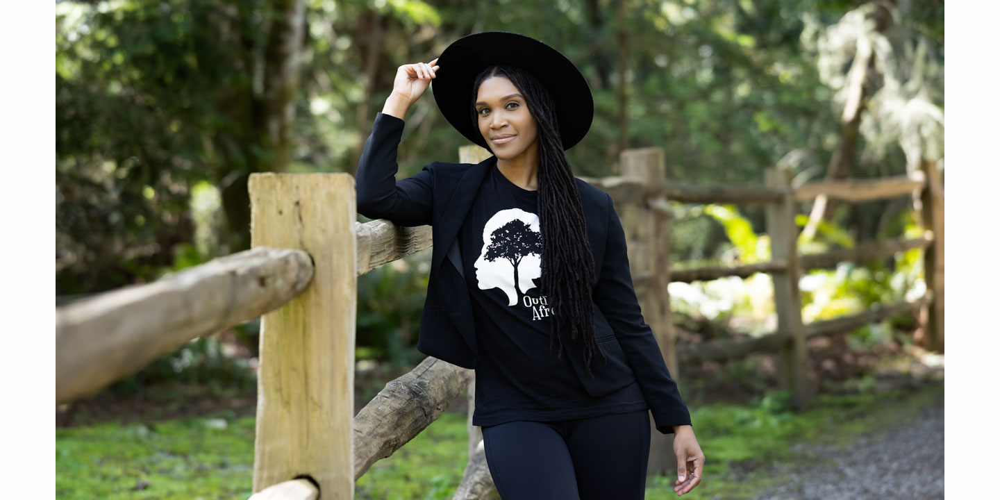 Mujer vestida con una camisa negra y un sombrero negro apoyada contra una valla con la mano en el sombrero, posando.